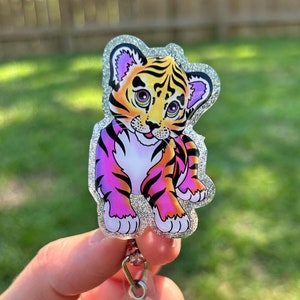 May include: A sparkly silver glitter badge reel with a cartoon tiger cub with pink and orange stripes. The tiger cub is smiling and has purple ears.