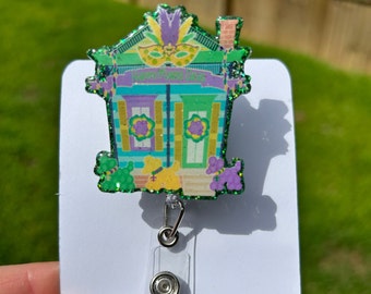 May include: A green, purple, and yellow glitter badge holder featuring a cartoon house with the text "Happy Mardi Gras".