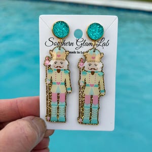 May include: A pair of gold glitter nutcracker earrings with turquoise glitter accents. The earrings are on a white card with the text "Southern Glam Lab made in Louisiana".