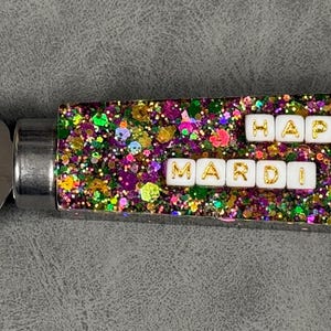 May include: A stainless steel cheese knife with a glittery purple, green, and gold handle. The handle has white tiles with gold lettering that reads "Happy Mardi Gras".