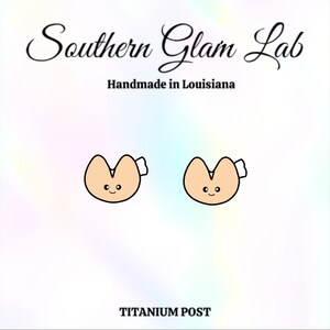 May include: A pair of gold-colored fortune cookie earrings with a smiling face. The earrings are made with a titanium post. The text "Southern Glam Lab" and "Handmade in Louisiana" is visible at the top of the image.