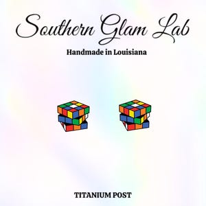 May include: A pair of colorful Rubik's Cube earrings. The earrings are made with a titanium post. The text "Southern Glam Lab" and "Handmade in Louisiana" are displayed at the top. The text "TITANIUM POST" is displayed at the bottom.
