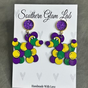 May include: Purple, green, and yellow balloon animal earrings with a purple glitter charm. The earrings are on a white card with the text "Southern Glam Lab" and "Handmade With Love".