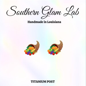 May include: A pair of earrings featuring a cornucopia filled with fall harvest fruits and vegetables. The earrings are made with a titanium post.