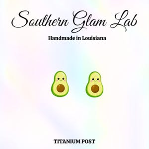 May include: A pair of avocado-shaped earrings with smiling faces. The earrings are green with a brown seed and are set against a white background. The text "Southern Glam Lab" and "Handmade in Louisiana" are at the top, and "TITANIUM POST" is at the bottom.