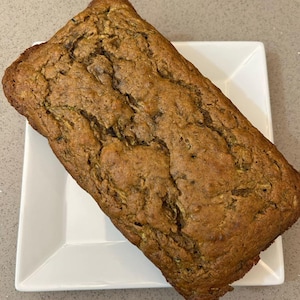 May include: A loaf of zucchini bread on a white square plate. The bread is a light brown color and has a slightly crumbly texture.