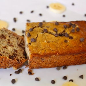 May include: A loaf of banana bread with chocolate chips, sliced and ready to eat. The bread is on a white plate with scattered chocolate chips and banana slices.