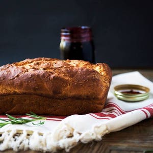 May include: A loaf of freshly baked bread sits on a white and red striped kitchen towel with a sprig of rosemary.