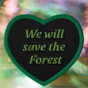 May include: A heart-shaped embroidered patch with a black background and a green border. The text "We will save the Forest" is stitched in green thread across the center of the patch in a bold, slightly slanted, sans-serif typeface.