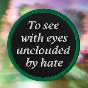 May include: A circular embroidered patch with a black background and a green border. The patch features the white embroidered text "To see with eyes unclouded by hate" arranged in four lines across the center of the circular fabric surface.