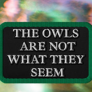 May include: A rectangular embroidered patch with a black background and a thick green border. The text THE OWLS ARE NOT WHAT THEY SEEM is stitched in white capital letters across the center of the patch in a bold, sans-serif font style.