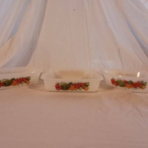 Puede incluir: Tres platos para hornear de cerámica blanca con un diseño floral que presenta verduras rojas, naranjas, amarillas y verdes. Los platos están dispuestos en una fila sobre una superficie blanca.