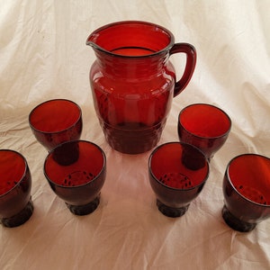 May include: A set of seven ruby red glass pieces, including a pitcher and six tumblers. The pitcher and tumblers have a textured, ribbed design.