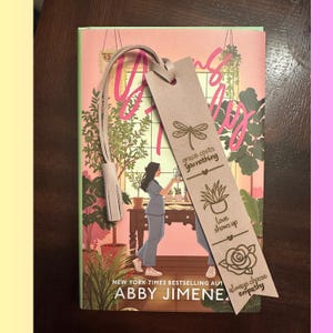 Könnte beinhalten: Ein Buch mit rosa und grünem Einband und einem hellrosa Lesezeichen. Das Lesezeichen hat eingravierte Designs und Text, darunter "grace costs you nothing" und "always choose empathy".