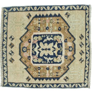 May include: A small, square rug with a beige background and a blue, brown, and pink geometric pattern. The rug has a fringed edge.