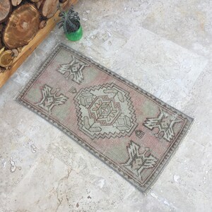 May include: A small, rectangular rug with a traditional Turkish kilim design. The rug is in shades of pink, beige, and gray. It has a geometric pattern with a central diamond shape.