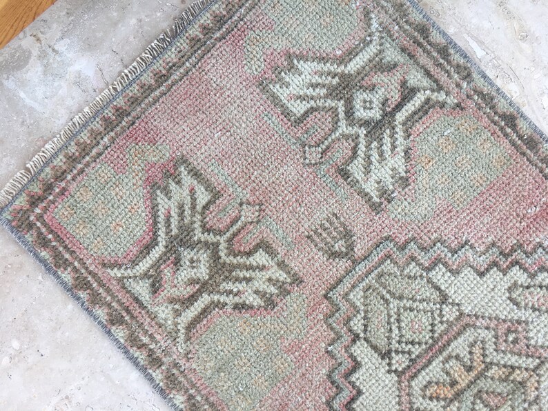 May include: A small, rectangular rug with a pink and gray color scheme. The rug has a traditional design with intricate patterns and a fringed edge.