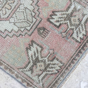 May include: A vintage rug with a faded pink background and a geometric pattern in shades of brown and gray. The rug has a fringed edge.
