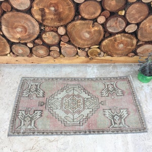 May include: A small, rectangular rug with a faded pink and cream color scheme. The rug has a traditional Turkish design with a central medallion and repeating patterns. The rug is lying on a light gray concrete floor.