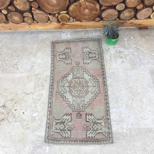 May include: A small, rectangular rug with a traditional Turkish design. The rug is in shades of pink, beige, and gray. It has a fringe border and is laid out on a light gray tiled floor.