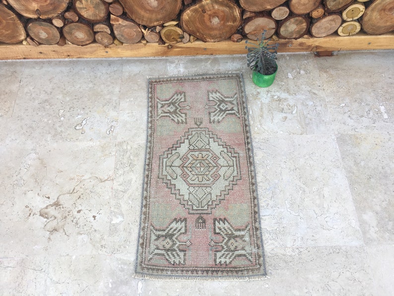 May include: A small, rectangular rug with a traditional Turkish design. The rug is in shades of pink, beige, and gray. It has a fringe border and is laid out on a light gray tiled floor.