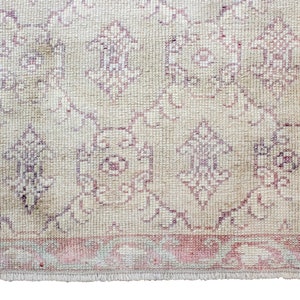 May include: A vintage rug with a cream background and a pink and purple floral pattern. The rug has a fringed edge.