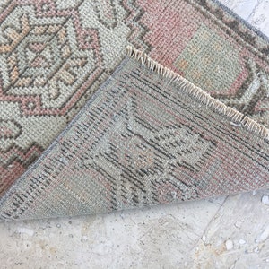 May include: A close-up of the back side of a light pink and gray patterned rug with a fringe border. The rug is folded in half, revealing the woven texture.