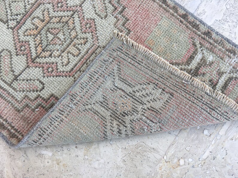 May include: A close-up of the back side of a light pink and gray patterned rug with a fringe border. The rug is folded in half, revealing the woven texture.
