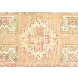 May include: A small, rectangular rug with a light pink background and a central cream-colored design. The design features a geometric pattern with pink, green, and cream accents.