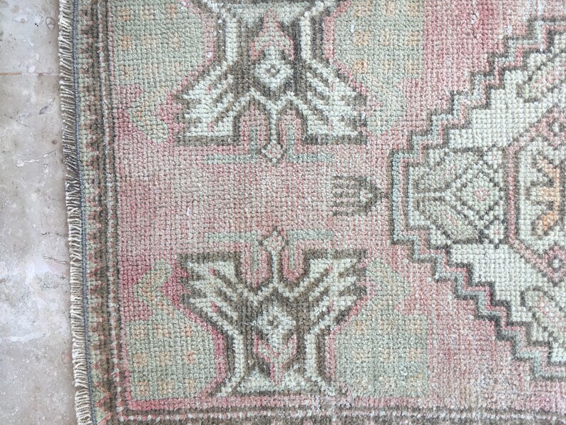 May include: A close-up of a pink and cream colored rug with a geometric pattern. The rug has a fringe border.