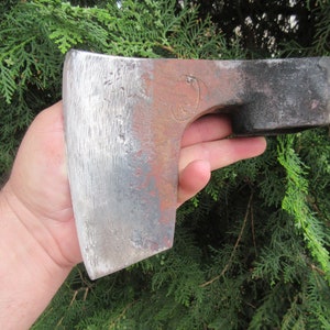 Puede incluir: Una mano sostiene un hacha grande, oxidada, de metal con un mango de madera. La cabeza del hacha es de color gris oscuro con una hoja gris claro. El hacha mide aproximadamente 45 cm de largo.