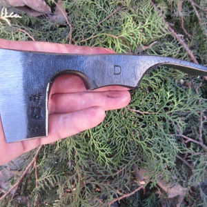 May include: A hand holds a small, black, curved axe with a D stamped on the handle. The axe has a sharp blade and a slightly curved handle.