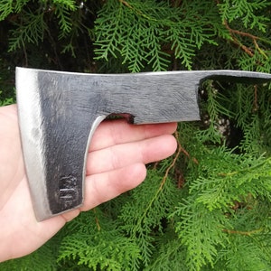 Könnte beinhalten: Eine schwarze Handaxt mit einer leicht gebogenen Klinge. Die Axt hat ein kleines Loch in der Klinge in der Nähe des Stiels. Die Axt ist aus Metall und hat ein raues, unfertiges Aussehen.