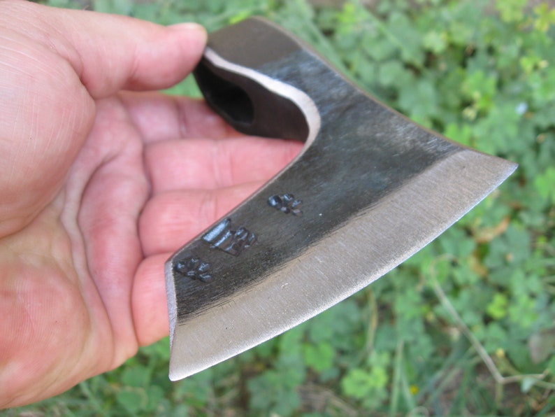 May include: A small, hand-forged axe with a black handle and a silver blade. The blade has a slight curve and is sharpened on one side. The axe is held in a hand, and the background is blurred.
