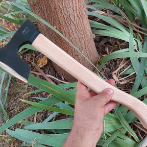 May include: A hand axe featuring a matte black metal head with the letters MF embossed on the side and a smooth, light-colored wooden handle. The axe head has a curved cutting edge and is securely attached to the straight, ergonomic wooden grip.