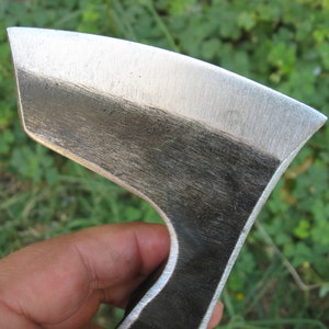 May include: Close-up of a hand holding a small, hand-forged axe with a polished steel head and a dark, unfinished wooden handle.