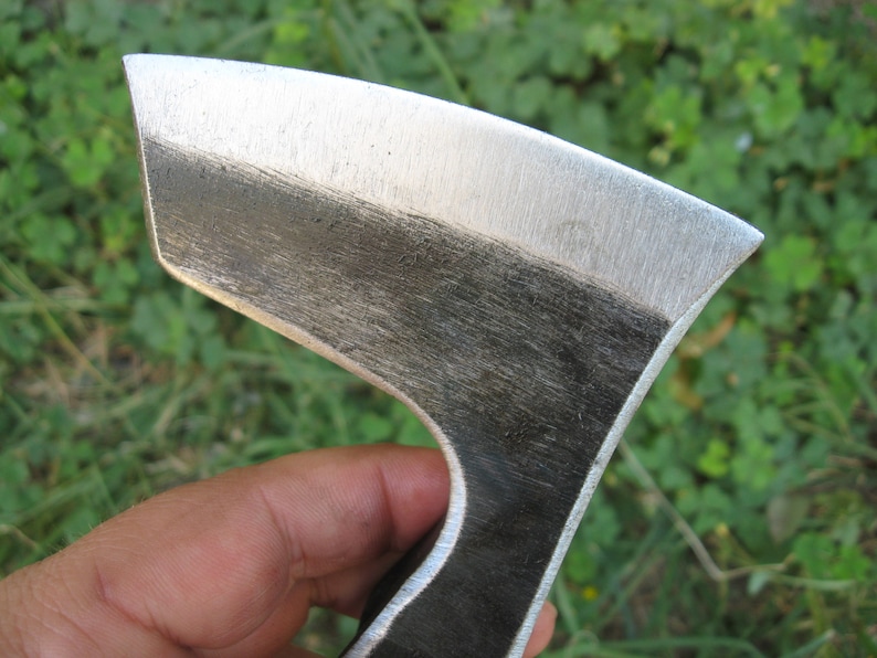 May include: Close-up of a hand holding a small, hand-forged axe with a polished steel head and a dark, unfinished wooden handle.