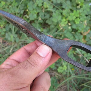 May include: A small, hand-forged axe head with a hole in the top for a handle. The axe head is made of steel and has a dark gray finish.