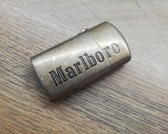 May include: A vintage-style, metallic lighter with the word "Marlboro" engraved on its surface. The lighter has a rectangular shape with rounded edges and a hinged lid. The lighter is made of a metal with a worn, antique finish.