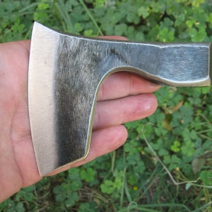 May include: A hand holding a small, hand-forged axe with a slightly curved blade. The axe is made of metal and has a dark gray finish.