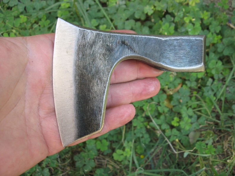 May include: A hand holding a small, hand-forged axe with a slightly curved blade. The axe is made of metal and has a dark gray finish.