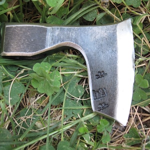 May include: A small, hand-forged axe with a curved blade and a wooden handle. The axe has a silver finish and is lying on a bed of green grass.
