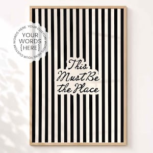 May include: A framed vertical wall art print featuring a pattern of thin black and white vertical stripes. The centre displays the text "This Must Be the Place" in a black script font. A circular badge in the top left corner reads "YOUR WORDS {HERE}".