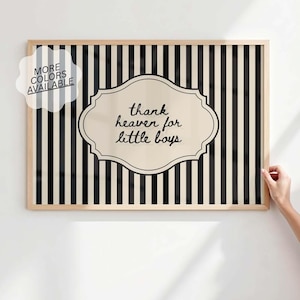 May include: Framed wall art with a black and white striped background. A cream-colored, ornate shape in the center contains the text "thank heaven for little boys" in black script. A small white bubble with the text "MORE COLORS AVAILABLE" is in the upper left corner.