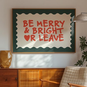 以下が含まれることがあります： 木製のキャビネット、ベージュの椅子、そして「BE MERRY & BRIGHT OR LEAVE」という赤い文字が緑色の背景に書かれた額装されたプリントが置かれた、ミッドセンチュリーモダンなリビングルームです。