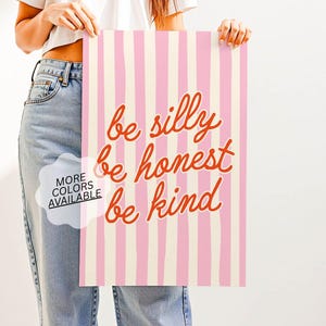 Op de afbeelding: Een roze en crèmekleurige gestreepte kunstprint met de woorden "be silly, be honest, be kind" in oranje script. De print wordt vastgehouden door een persoon die een wit t-shirt en een blauwe jeans draagt. De woorden "MORE COLORS AVAILABLE" zijn ook zichtbaar.