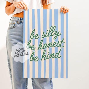 Op de afbeelding: Een rechthoekige poster met verticale strepen in lichtblauw en beige. De poster bevat de woorden "be silly, be honest, be kind" in groene cursieve letters. Een witte tekstballon met de tekst "MORE COLORS AVAILABLE" staat linksonder.