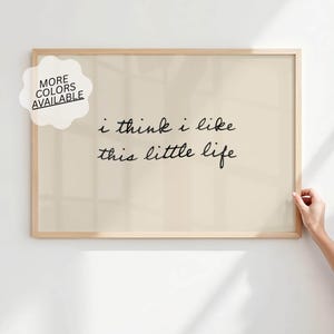 Puede incluir: Una impresión enmarcada con el texto "i think i like this little life" en escritura negra. La impresión tiene un fondo beige claro y un marco de madera natural. Una pegatina en la esquina superior izquierda dice "MORE COLORS AVAILABLE".