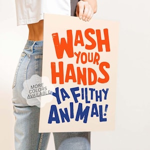 May include: A cream-colored poster with the words "WASH YOUR HANDS YA FILTHY ANIMAL!" in orange and blue block letters. The poster is held by a person wearing a white shirt and blue jeans. A small graphic says "MORE COLORS AVAILABLE".