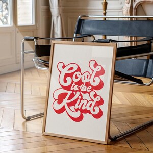 Puede incluir: Una impresi&oacute;n rectangular enmarcada que presenta el texto Cool to be Kind en una fuente retro roja y audaz con un contorno blanco. La obra de arte se muestra en un marco de madera de color claro, exhibiendo un dise&ntilde;o limpio y tipograf&iacute;a.